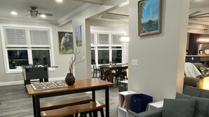 Ein geräumiges Wohnzimmer mit grauem Sofa, einem Holzesstisch und Bänken. Im Hintergrund befindet sich eine Küchenzeile mit Kühlschrank und Spüle.