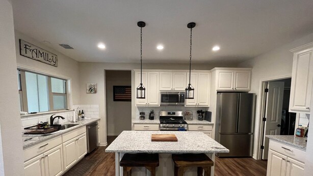 A modern kitchen with white cabinets, a central island with stools, stainless steel appliances, and pendant lighting. - Charming new home near Canyon Lake/Guadalupe River (Canyon Lake)