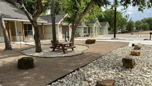 A patio area with a picnic table surrounded by gravel and large rocks, trees, and a building with a sloped roof in the background. - Charming new home near Canyon Lake/Guadalupe River (Canyon Lake)