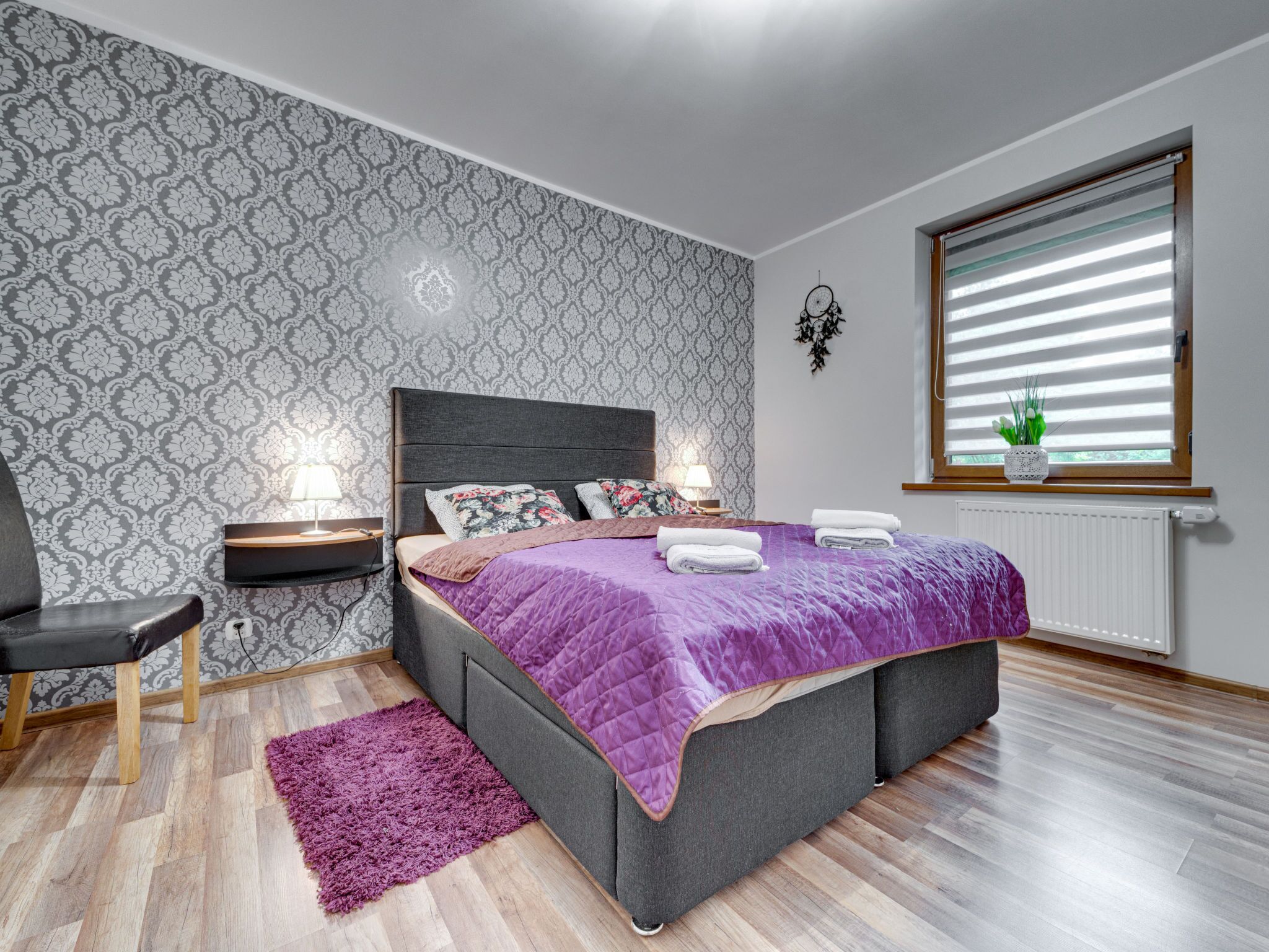 A modern bedroom with a large bed, a nightstand, a chair, and a window with blinds.