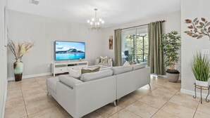 Una sala de estar moderna con un sofá blanco, un televisor de pantalla plana, una araña y un ventanal con cortinas.