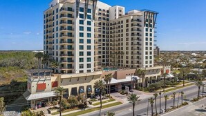 A multi-story hotel with a modern design, featuring balconies on each floor, a rooftop pool, and a landscaped front area with palm trees.