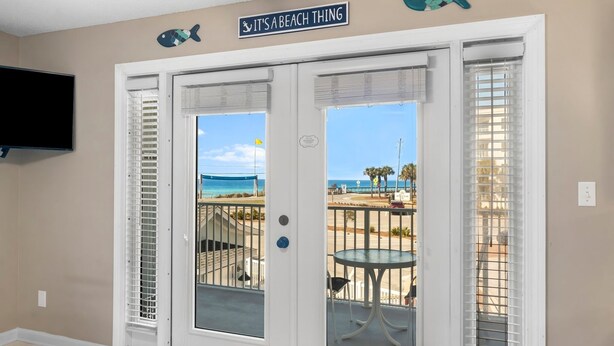 A room with a view of the beach, a sign above the door reading "IT'S A BEACH THING", and a table with chairs on the balcony.