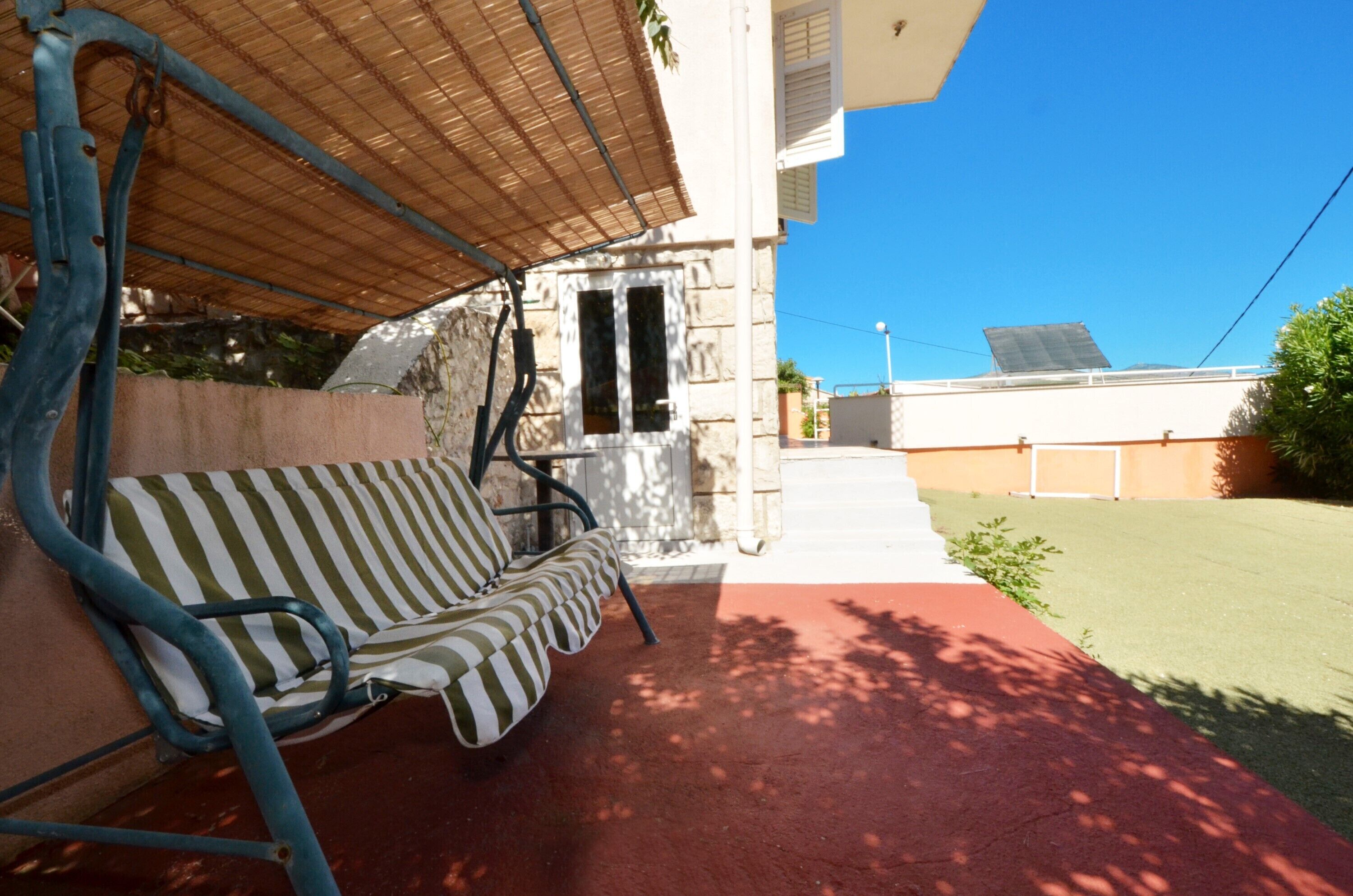 A covered outdoor seating area with a striped sofa, a metal chair, and a red pathway leading to a building entrance.