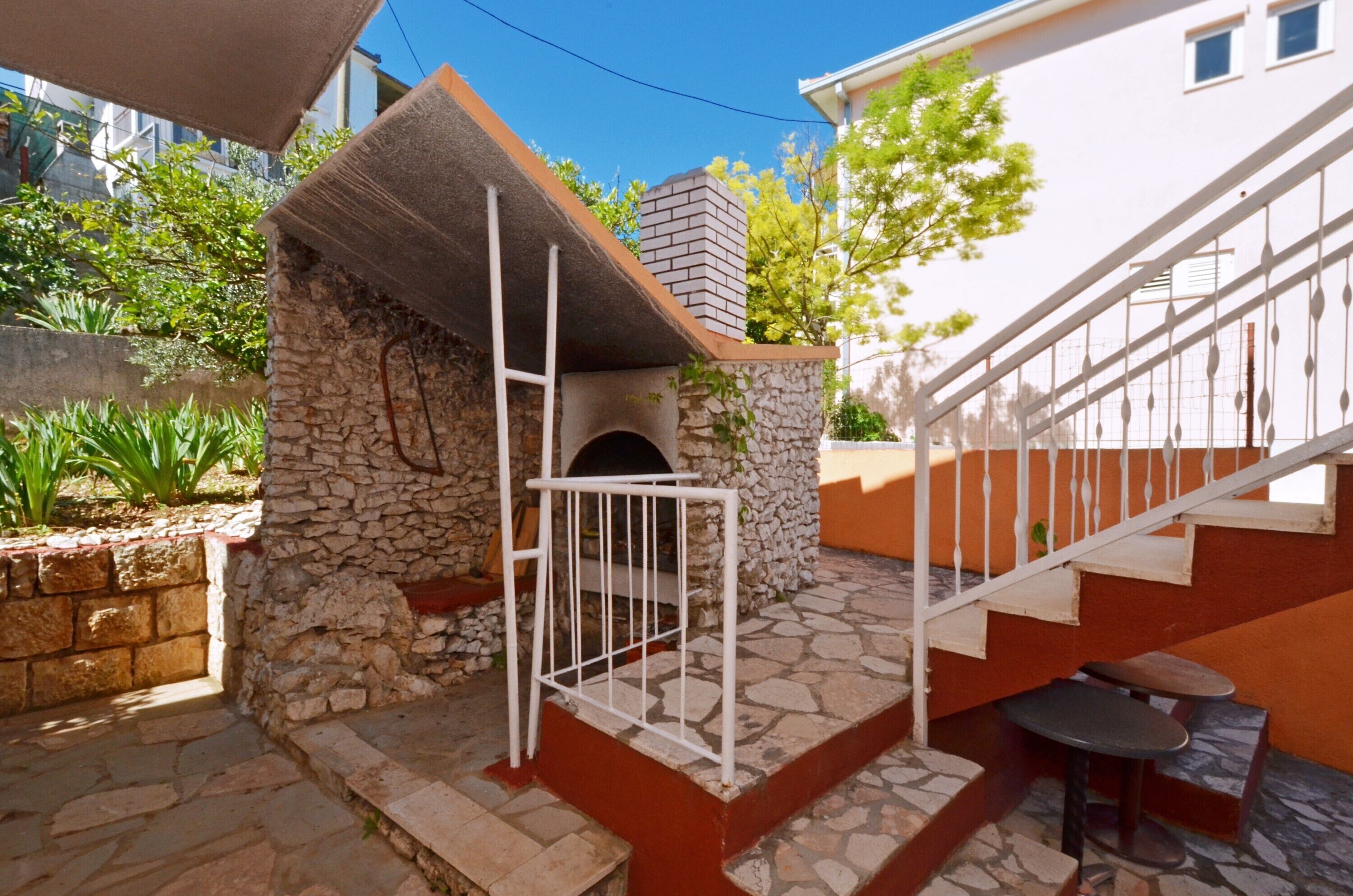 A stone-built outdoor area with a small brick oven, a white metal gate, and a staircase leading to a building with orange walls.