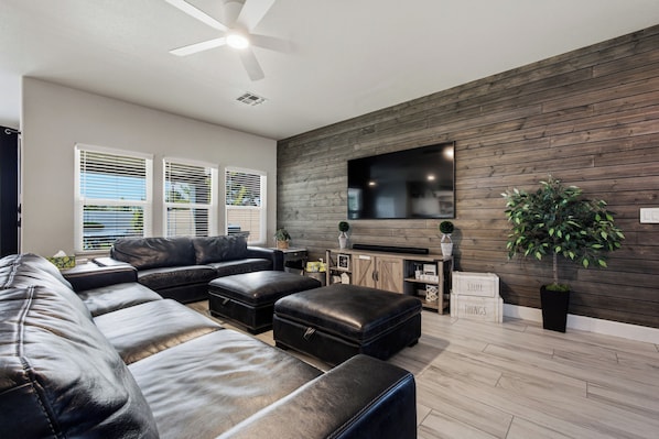 A modern living room with a sectional sofa, a flat-screen TV mounted on a wooden wall, and a ceiling fan.