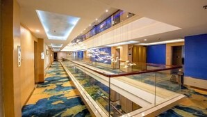 Pasillo de hotel con barandilla de vidrio, alfombra estampada e iluminación empotrada en el techo.