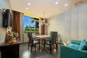 Un área de comedor con una mesa y sillas, un sofá, un televisor montado en la pared y un ventanal con vistas a la vegetación.