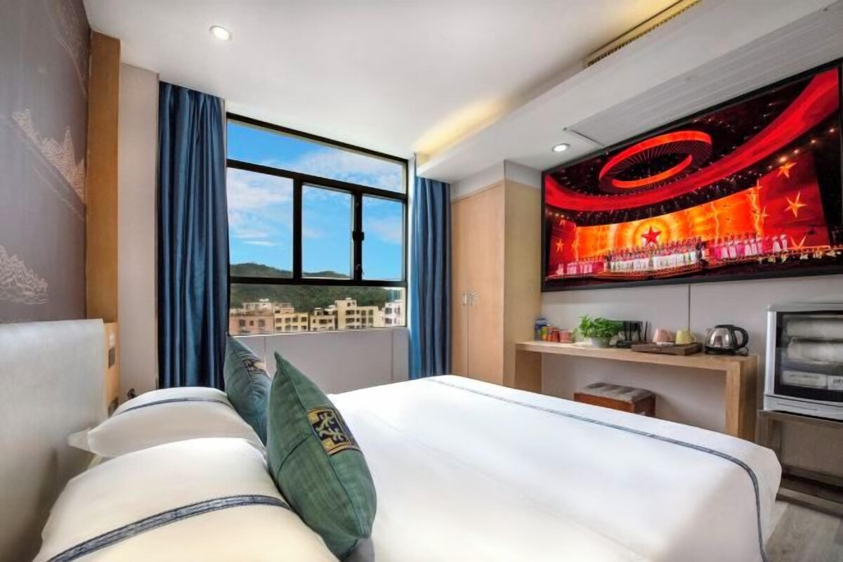 A hotel room with a bed, a window with a city view, a wall-mounted TV, and a shelf with items.
