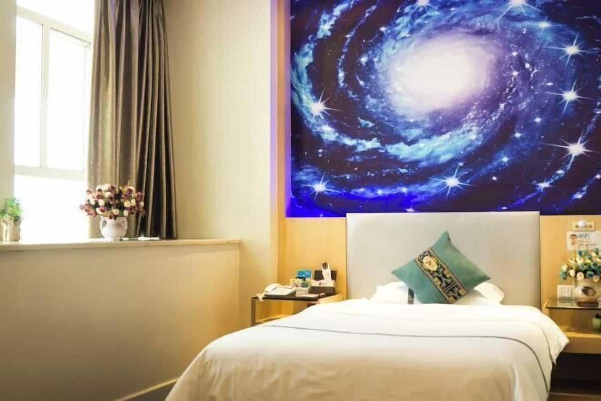 Ein Hotelzimmer mit Bett, Nachttisch, Blumenvase und Wandgemälde einer Galaxie.