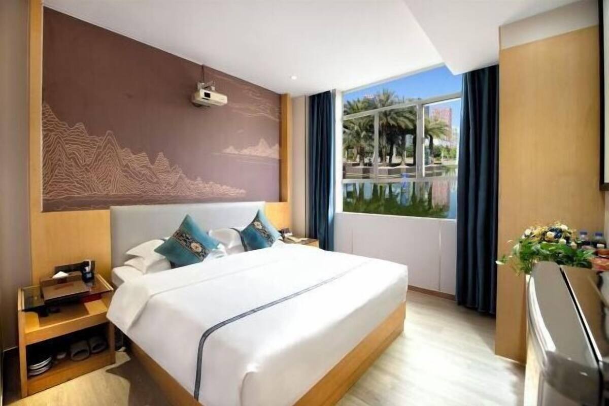 Ein Hotelzimmer mit einem großen Bett, einem Nachttisch, einem Schreibtisch und einem Fenster mit Blick auf Palmen.