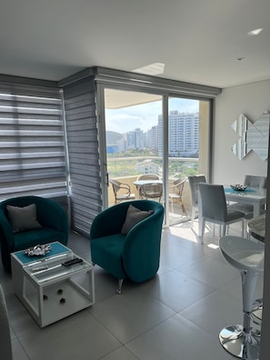 A modern living room with teal armchairs, a glass coffee table, and a dining area with a white table and chairs.