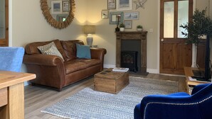 A cozy living room with a brown leather sofa, a wooden coffee table, and a blue armchair. A fireplace with framed pictures and a star decoration is visible. - Vineyard Cottage Sleeps 4 with Garden (Norwich)