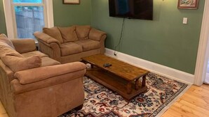 A living room with two beige sofas, a wooden coffee table, a flat-screen TV mounted on the wall, and a patterned rug on the floor. - Trip6 Historic Brick Home (Helena)
