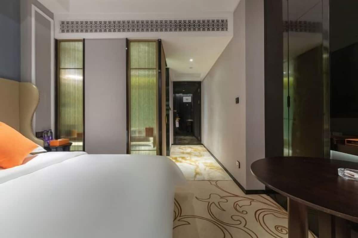 Une chambre d’hôtel avec un lit, une table de chevet, un miroir et une vue sur un couloir dont le sol est recouvert d’une moquette à motifs.