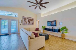 A spacious living room with a large sofa, a ceiling fan, a flat-screen TV, and a wooden floor. - Spacious Getaway ~ Heated LED Pool & Ping Pong (Minneola)