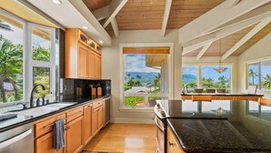 Une cuisine moderne avec des meubles en bois, un plan de travail noir et une vue sur les palmiers et les montagnes, visible à travers de grandes fenêtres.