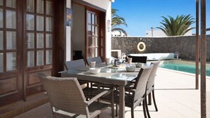 An outdoor dining area with wicker chairs and a glass table, a stone wall, and a pool in the background. - Villa Serendipity with Mountain View, Private Heated Pool & Wi-Fi (Playa Blanca)