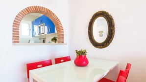 A dining area with a white table, red chairs, a red vase with a plant, a mirror with an ornate frame, and a brick archway leading to a kitchen.