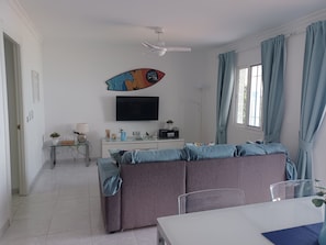 A modern living room with a sofa, a TV mounted on the wall, a dining table, and a ceiling fan.