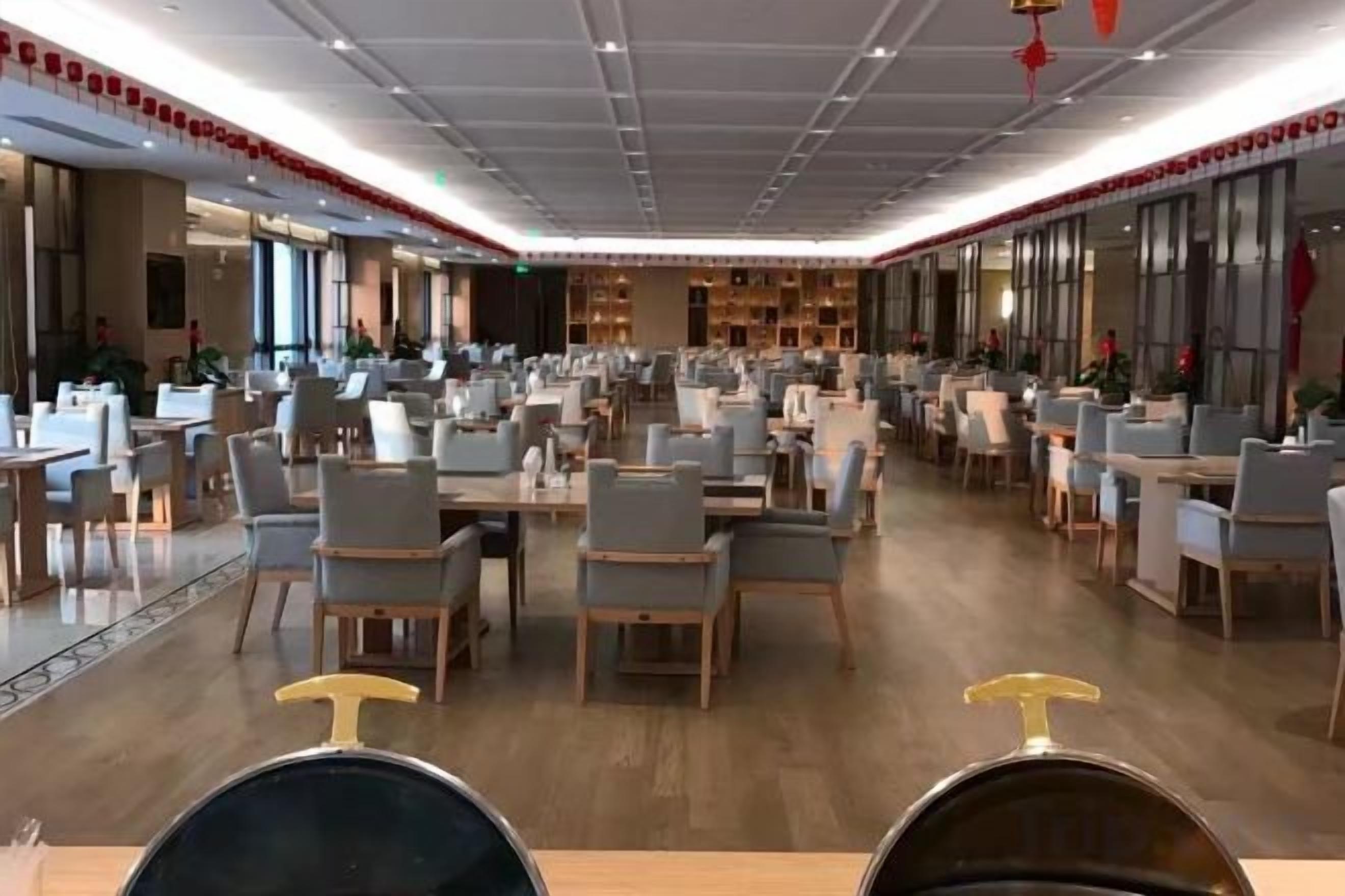 Uma sala de jantar ampla com várias mesas e cadeiras, teto alto com iluminação embutida e grandes janelas em um dos lados.