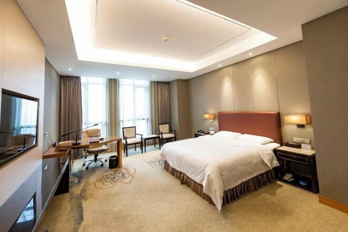 Ein Hotelzimmer mit einem großen Bett, einem Schreibtisch mit Stuhl, einem Fernseher und einem Fenster mit Vorhängen.
