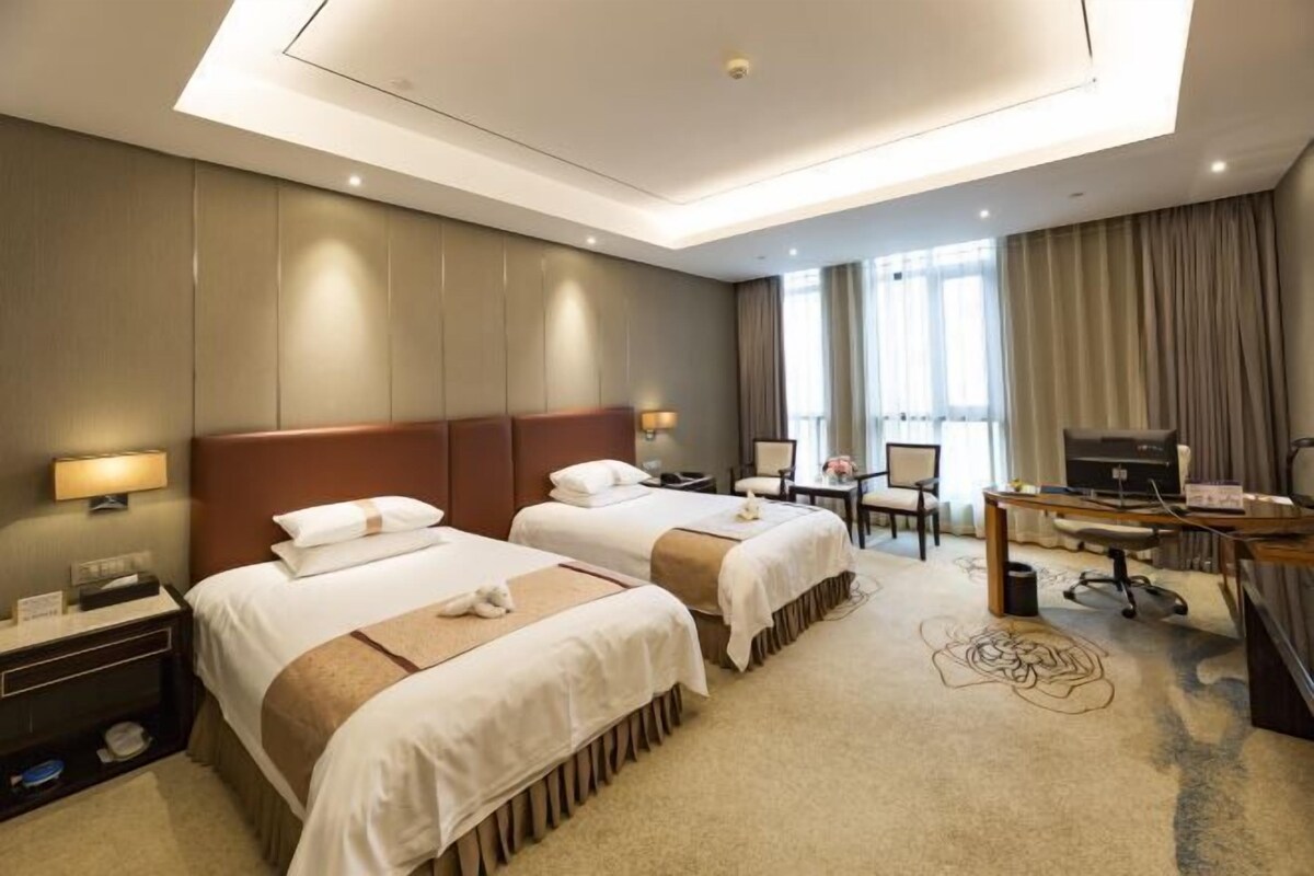 Ein Hotelzimmer mit zwei Betten, einem Schreibtisch, einem Stuhl, einem Fernseher und einem Fenster mit Vorhängen.