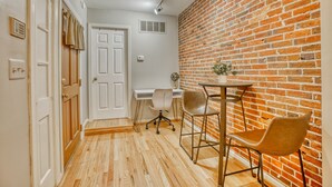 A room with a wooden floor, white walls, a brick wall, a desk with two chairs, and a door. - Charming Federal Hill! One Bedroom With Vibes (Baltimore)