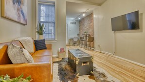 A living room with a leather sofa, a vintage trunk, and a flat-screen TV mounted on the wall. - Charming Federal Hill! One Bedroom With Vibes (Baltimore)