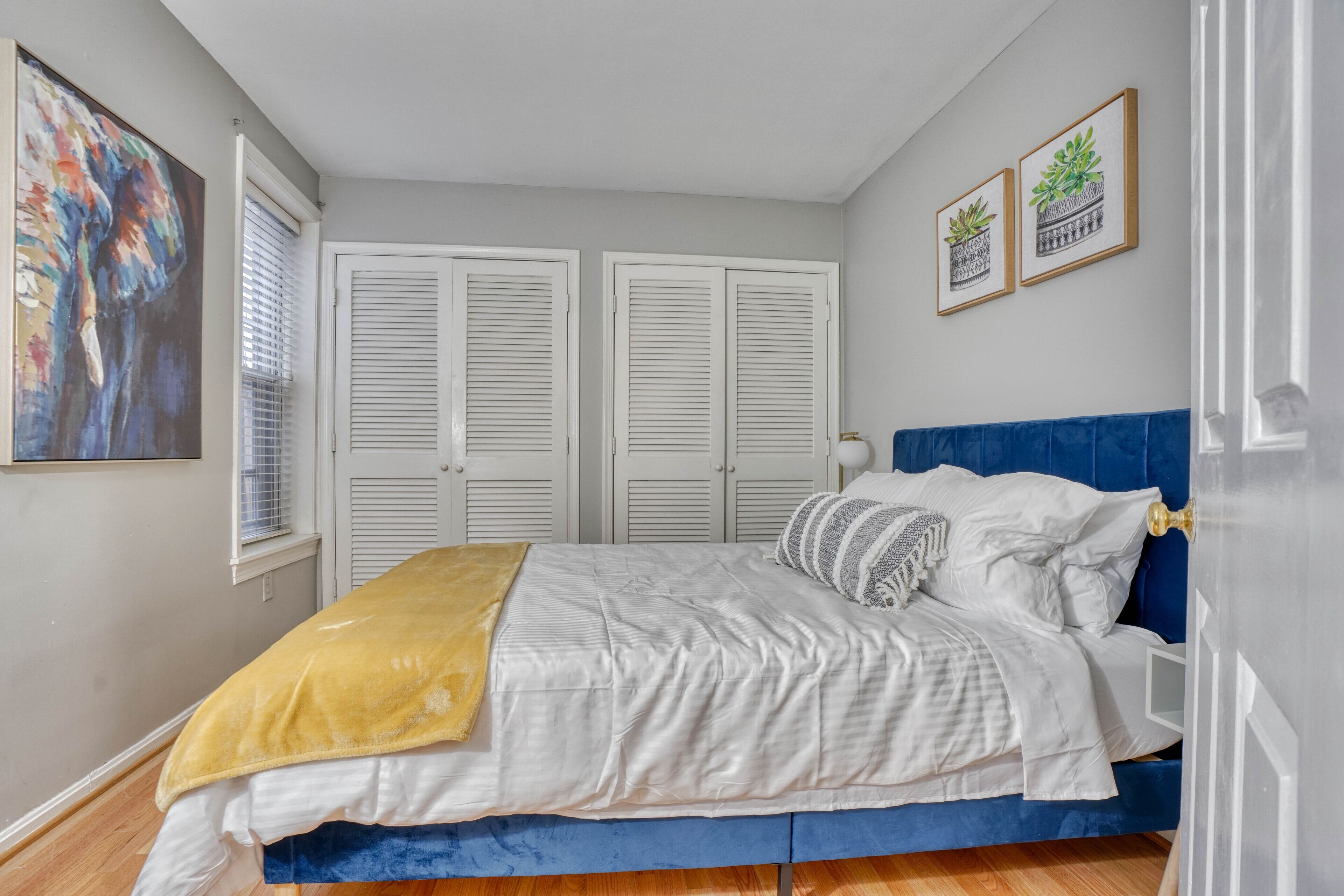 A bedroom with a bed, white and blue bedding, a yellow throw blanket, and a painting on the wall.
