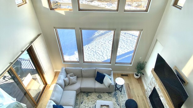 A spacious living area with a large sectional sofa, a coffee table, and a fireplace. The room features large windows offering a view of a snowy landscape and a building.