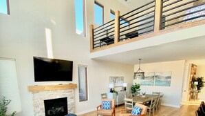 A spacious living room with a high ceiling, a fireplace, a dining area, and a staircase to an upper level.