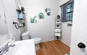 Une salle de bain avec un lavabo, des toilettes et une douche, le tout de couleur blanche. On y trouve des étagères murales ornées de plantes et de serviettes, ainsi qu’une fenêtre avec des rideaux.