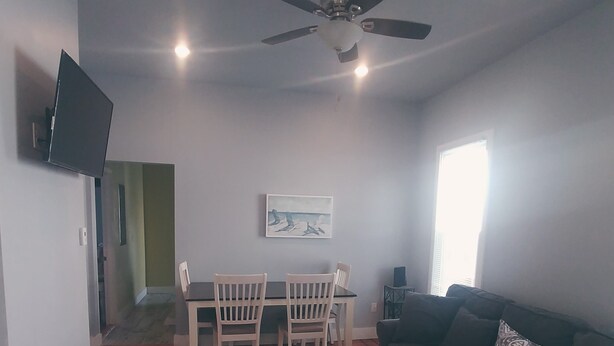 A living room with a wooden floor, a ceiling fan, a TV mounted on the wall, a dining table with chairs, a sofa, and a window. - Eastbayside/Munjoy - Charming Downtown Escape (Portland)