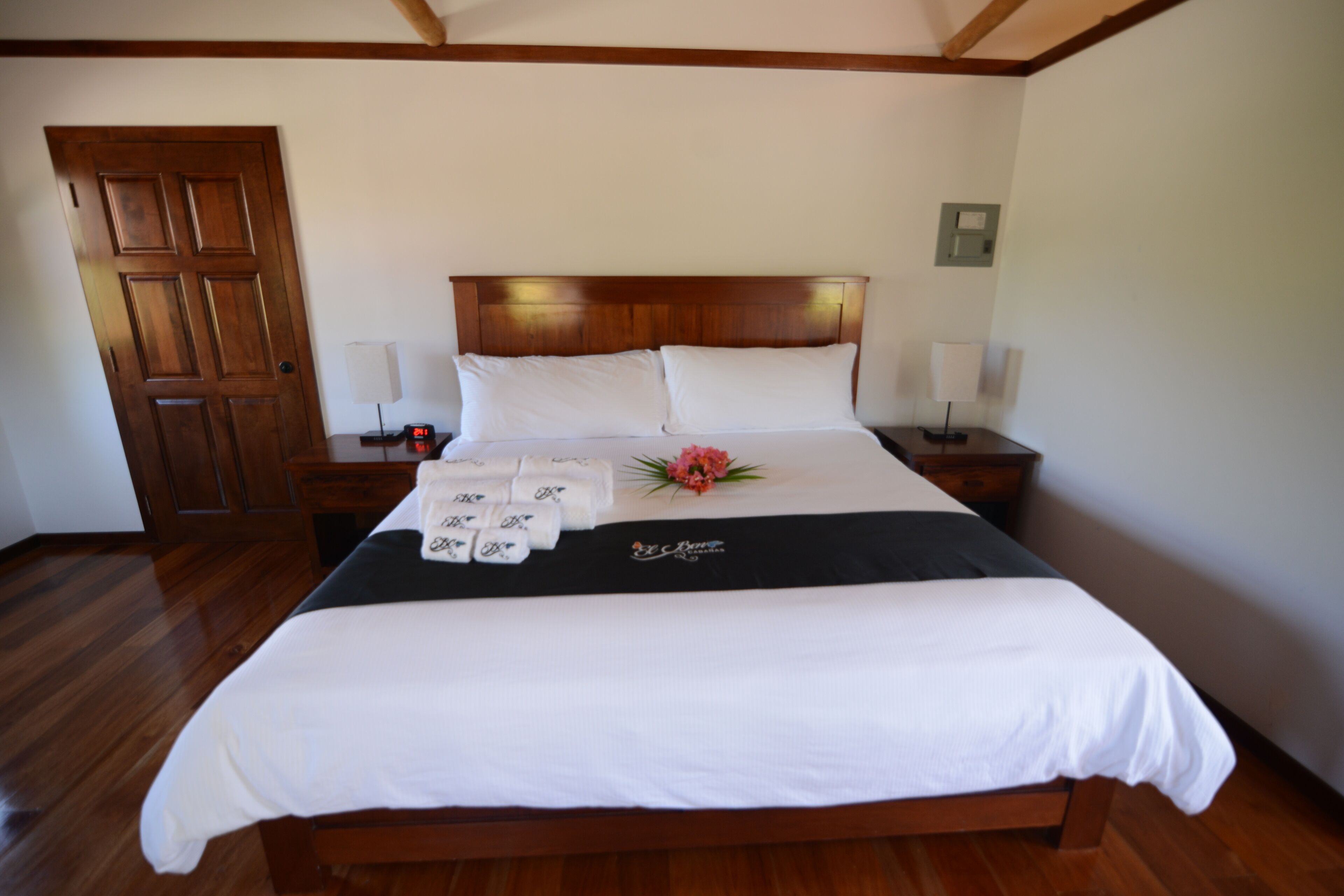 Una cama bien tendida con sábanas blancas y un cabecero negro, una puerta de madera y una mesita de noche con una lámpara y toallas.