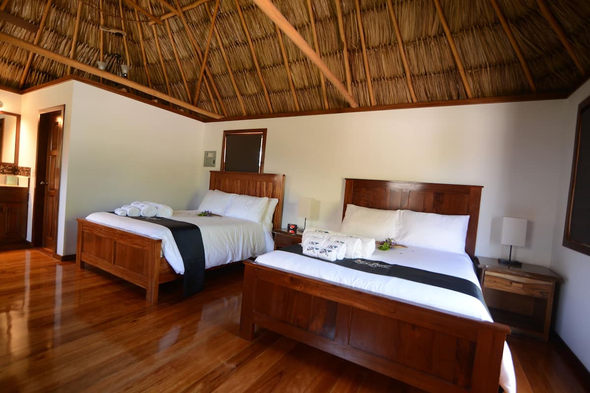 Una habitación de hotel con dos camas de madera, techo de paja, suelos de madera, una mesita de noche con lámpara y un espejo en la pared.