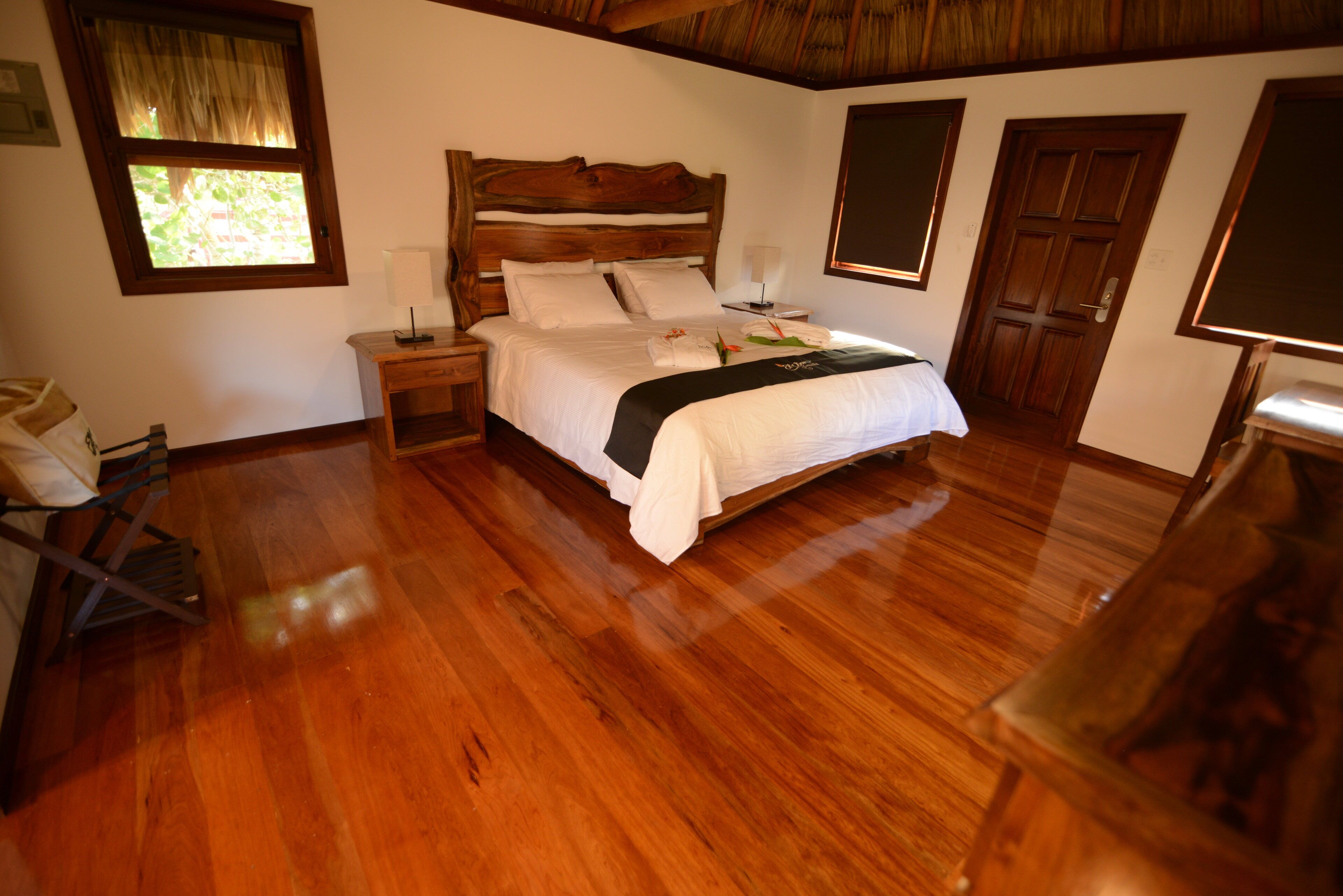 A spacious bedroom with a large wooden bed, wooden floors, a nightstand, and a window with curtains.