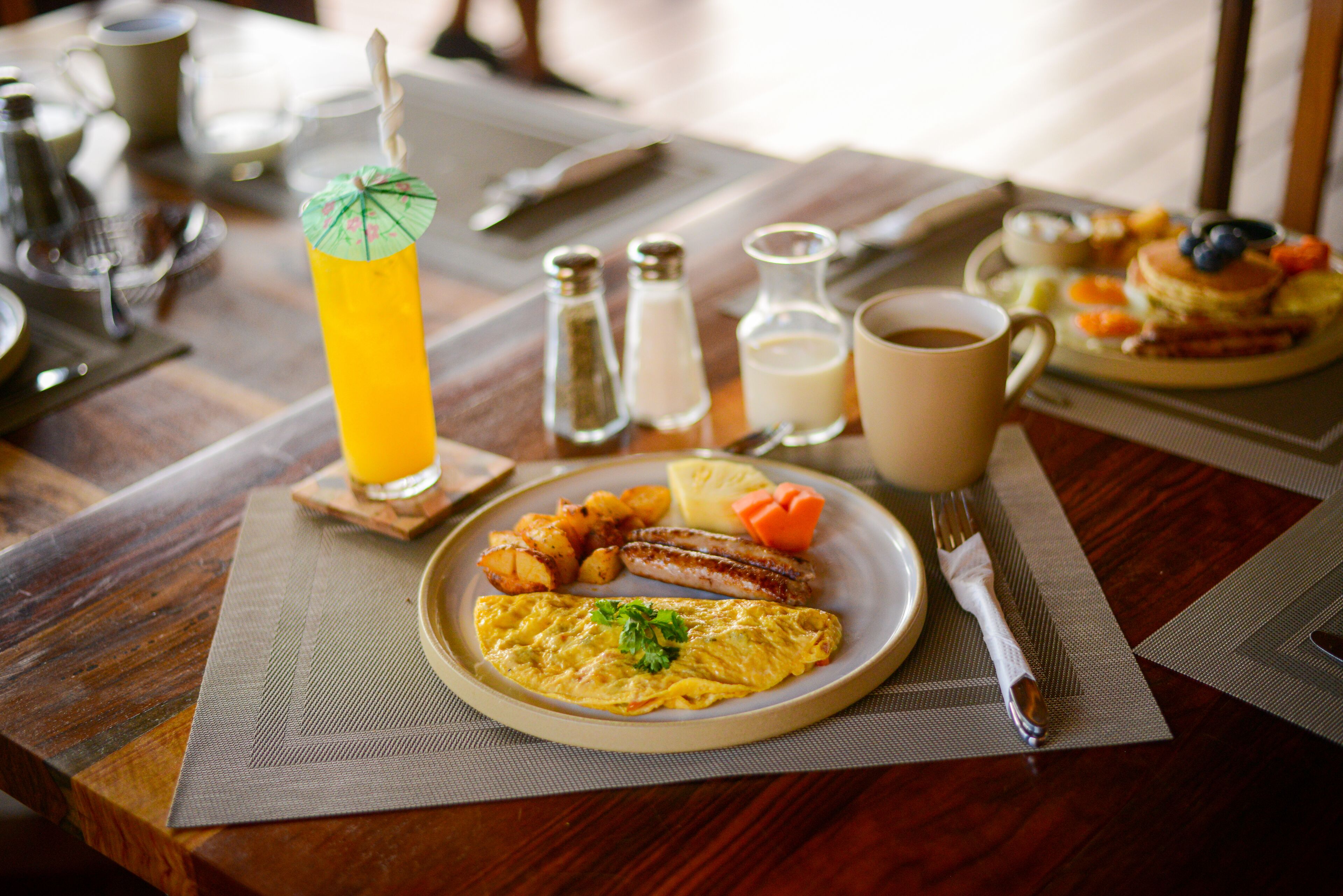 Une table en bois dressée pour le petit-déjeuner, avec une assiette d’œufs, de saucisses et de pommes de terre, un verre de jus d’orange et une tasse de café.