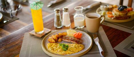 Une table en bois dressée pour le petit-déjeuner, avec une assiette d’œufs, de saucisses et de pommes de terre, un verre de jus d’orange et une tasse de café.