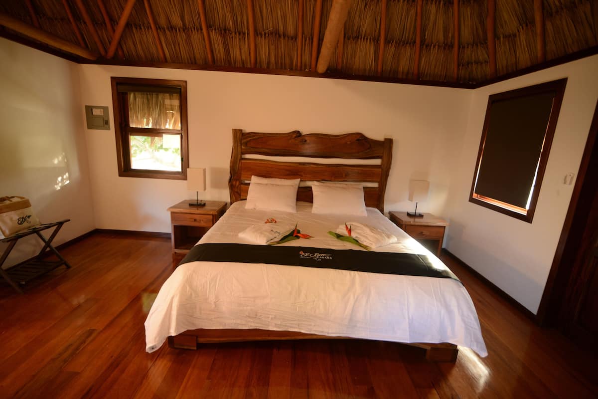 Una cama bien hecha con cabecera de madera, ropa de cama blanca y un arreglo decorativo de almohadas. Hay dos mesitas de noche con lámparas, una ventana con vistas y un suelo de madera.