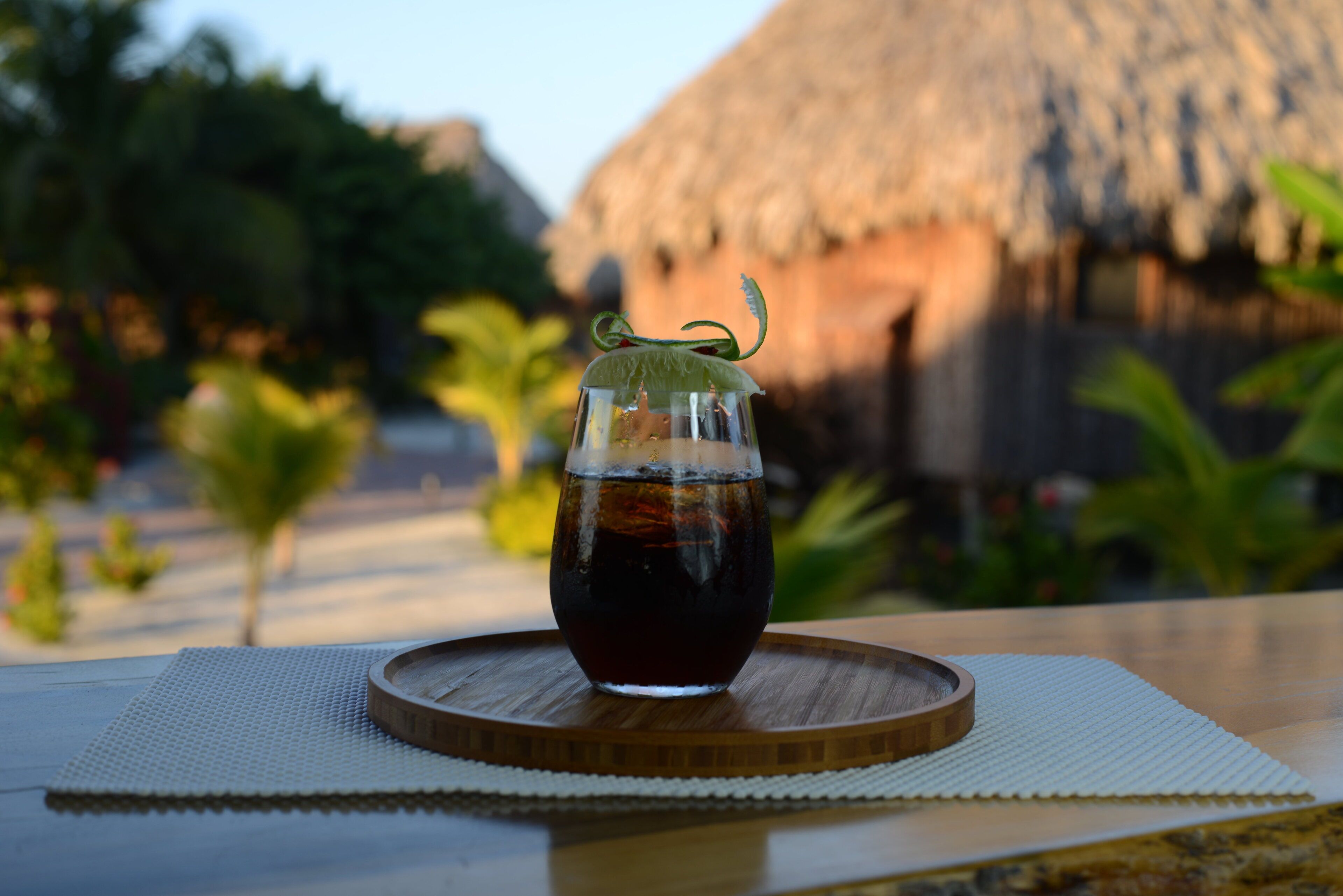 A glass of dark beverage on a wooden coaster with a thatched roof structure in the background.