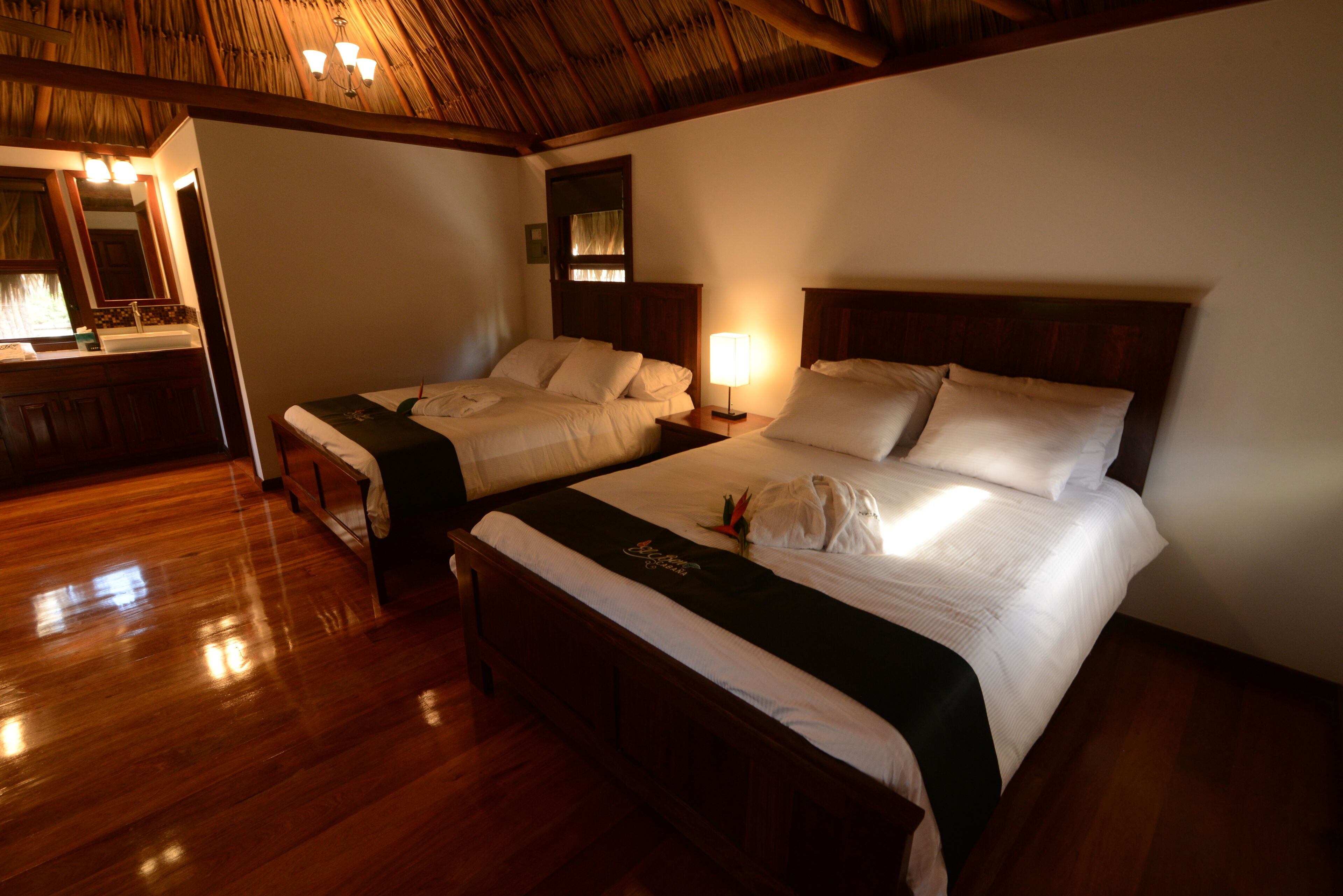 Habitación de hotel con dos camas, piso de madera, baño con espejo y lavamanos, y techo con vigas a la vista.