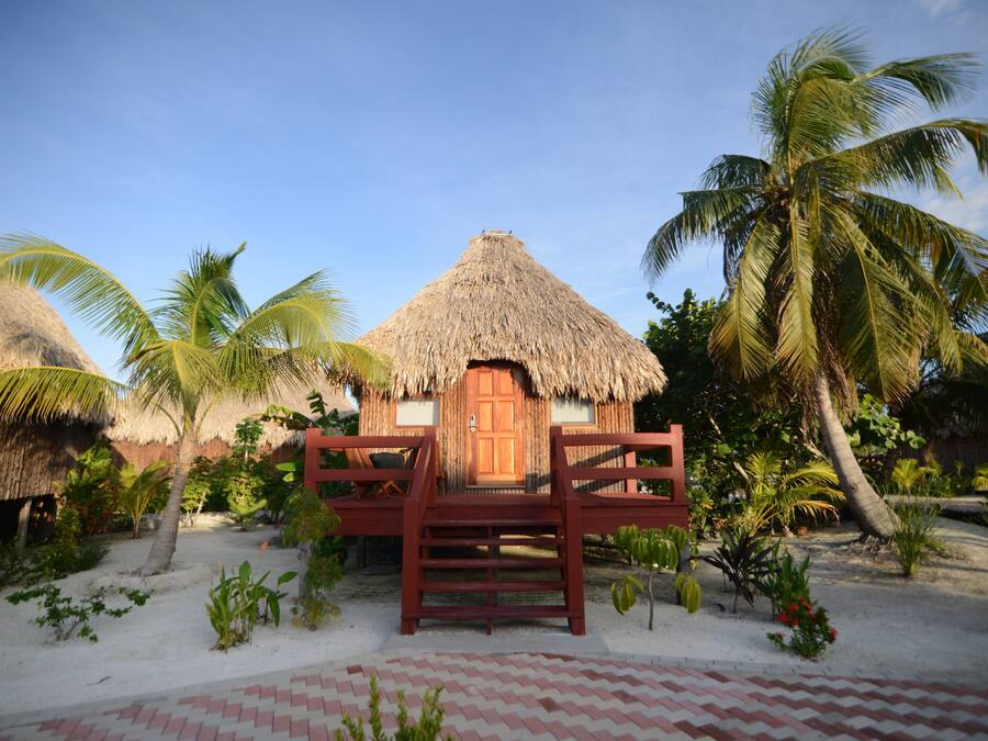 Ein reetgedecktes Bungalow mit Holztür und kleiner Veranda, umgeben von Palmen und einem Sandstrand.