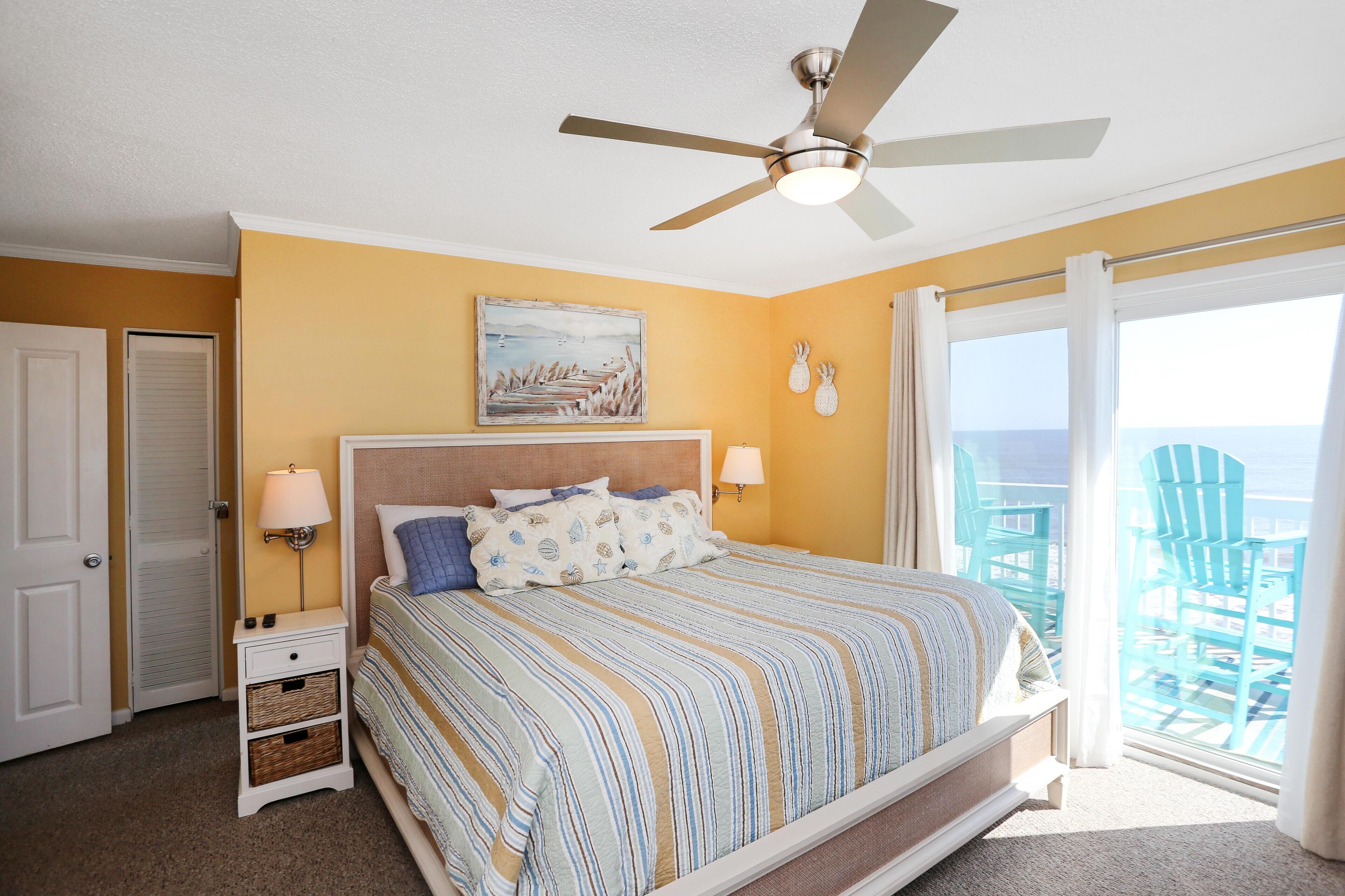 A bedroom with a bed, a ceiling fan, a painting, and a view of the outside.