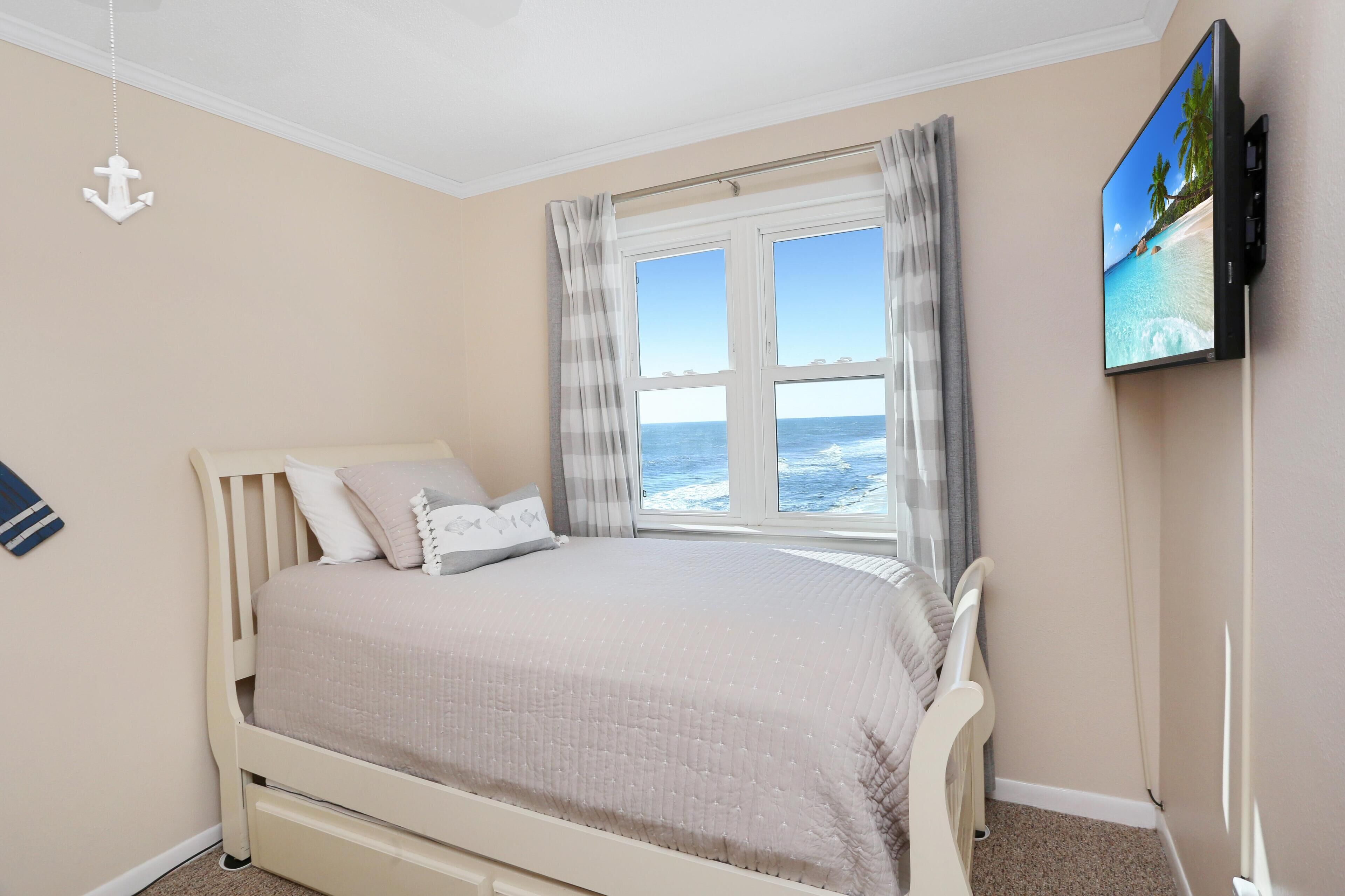 A bedroom with a bed, a TV on the wall, a window with curtains, and a view of the sea.