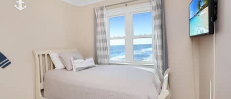 A bedroom with a bed, a TV on the wall, a window with curtains, and a view of the sea.