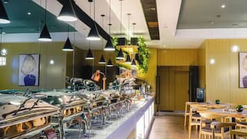 Uma área de buffet com várias estações de comida, luminárias pendentes e um espaço de estar com mesas e cadeiras de madeira.