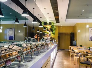 Un espace buffet comprenant plusieurs présentoirs de nourriture, des luminaires suspendus et un coin salon avec des tables et des chaises en bois.