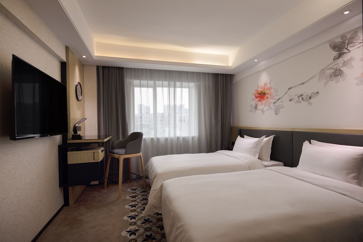 Ein Hotelzimmer mit zwei Betten, einem Schreibtisch mit Stuhl, einem Flachbildfernseher und einem großen Fenster mit Vorhängen.