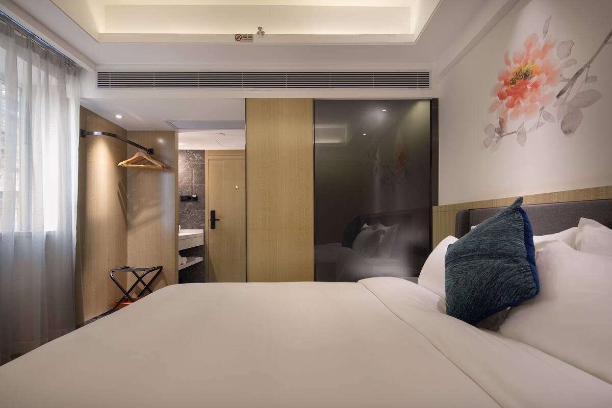 Une chambre d’hôtel moderne dotée d’un grand lit, d’un dressing, d’une salle de bain avec douche et d’une grande fenêtre avec rideaux.